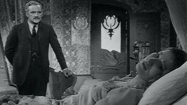 A black-and-white horror-film still showing a man standing beside someone lying in bed